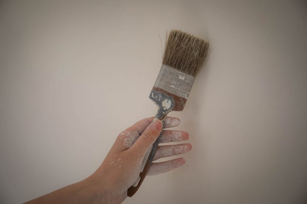 A hand holds a paintbrush with white paint against a plain wall, showcasing DIY home improvement.